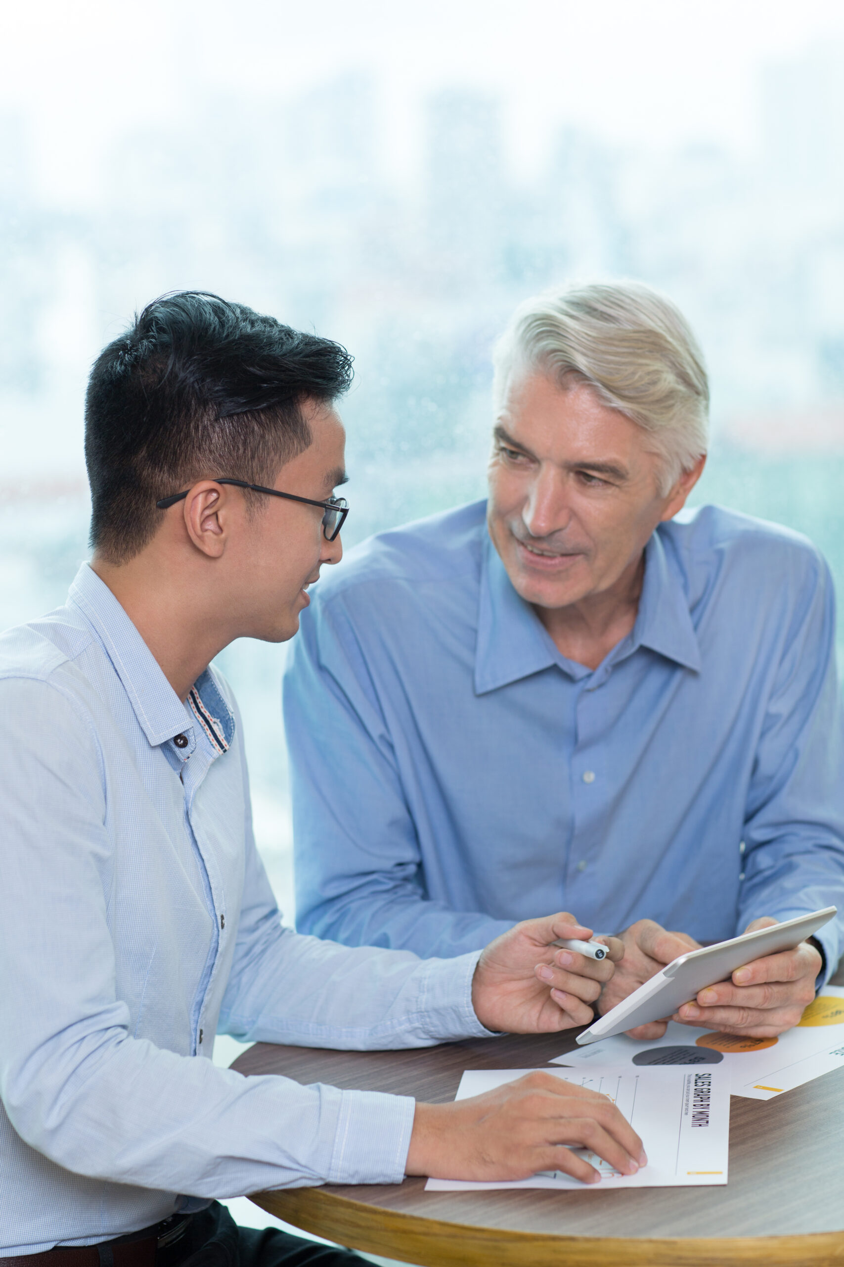 Business team using tablet when discussing project. Senior Caucasian businessman showing presentation on portable device, Asian colleague asking question. Teamwork or cooperation concept